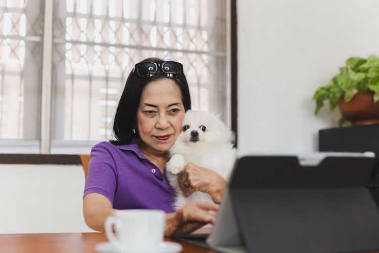 Senior woman holding pomeranian dog while working laptop