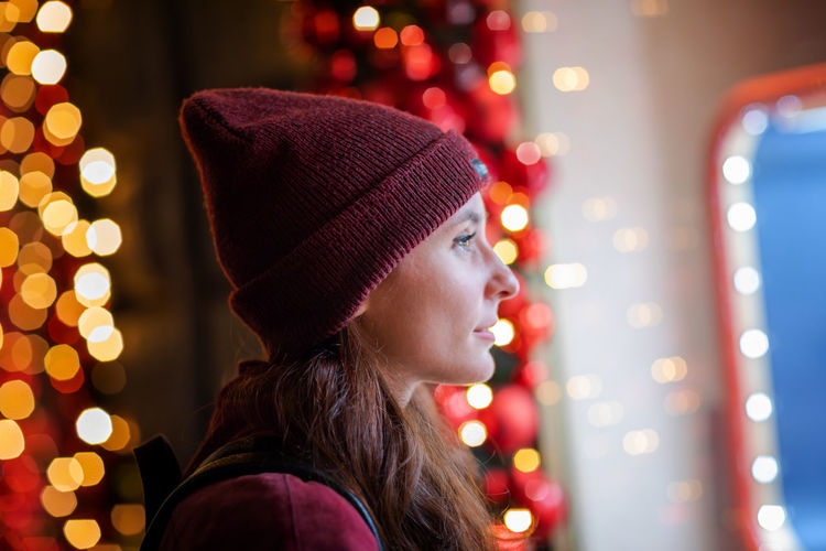 Young girl in a woolen hat looks at the shop windows decorated with christmas lights.