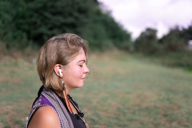 Side view of calm young woman with blond hair in casual clothes and wireless earphones listening to music and meditating with closed eyes in nature