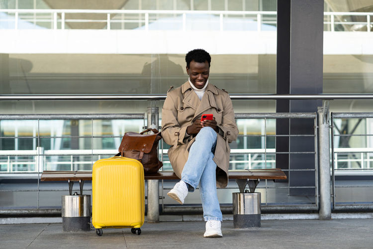 Black traveler man sitting on bench in airport terminal using mobile phone, waiting public transport