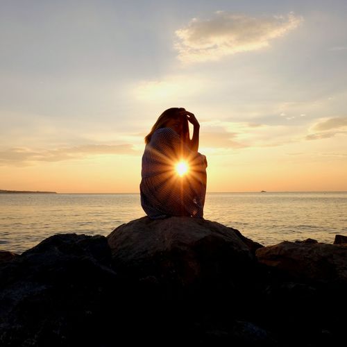 Side view of teenage girl sitting by sea against sky during sunset