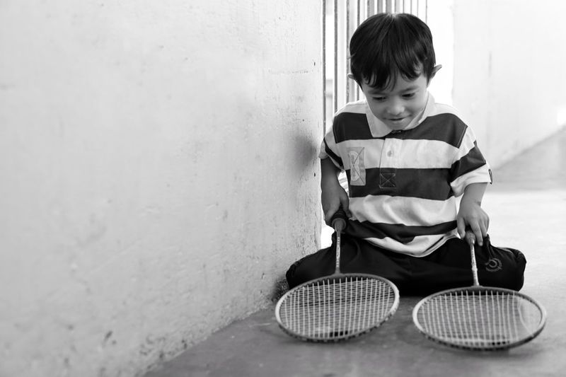 Boy holding badminton rackets while sitting by wall