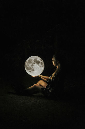 Optical illusion of woman holding moon while sitting on field at night