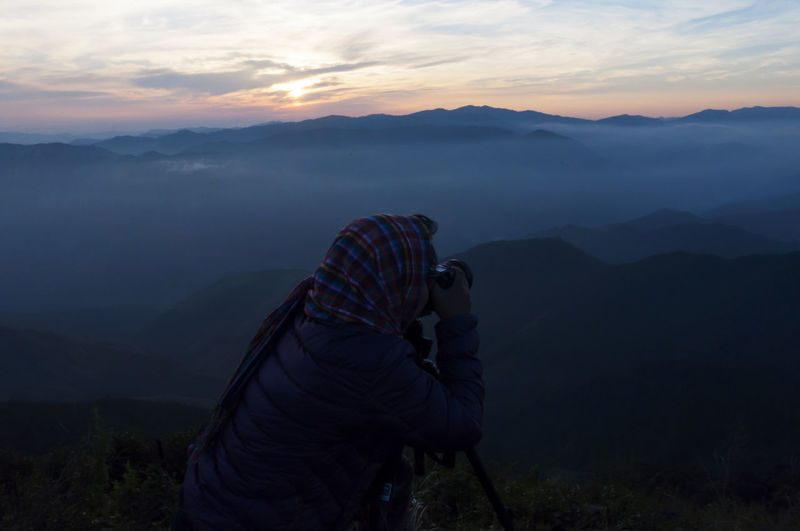 Rear view of woman photographing mountains against sky during sunset