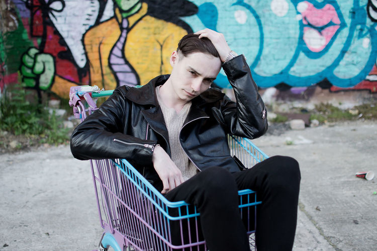 Young man sitting in shopping cart against graffiti on wall