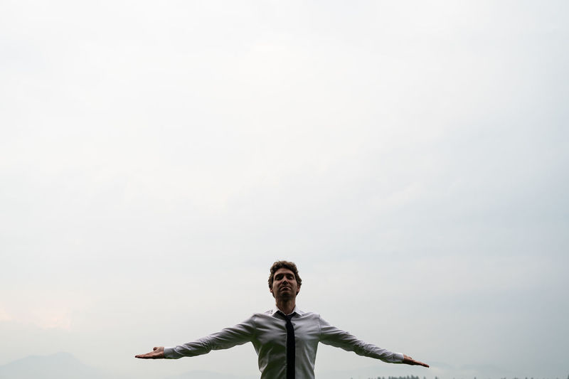 Portrait of young man with arms outstretched standing against sky