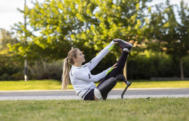 Side view of professional female runner with bionic prosthesis stretching legs during workout while sitting on grass