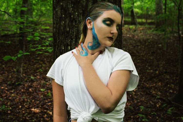 Young woman with face paint leaning on tree trunk in forest