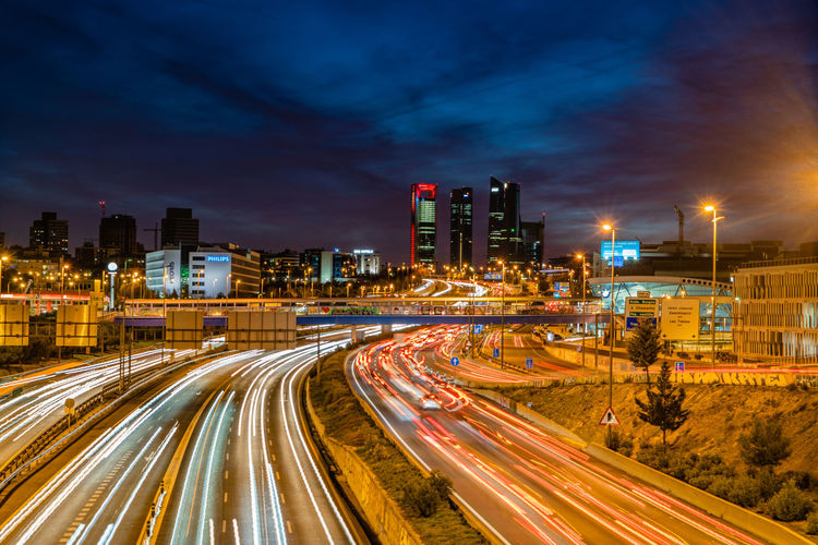 Light trails on road amidst buildings against sky at night