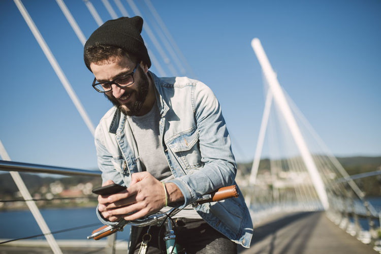 Smiling young man with fixie bike using a smartphone on a bridge