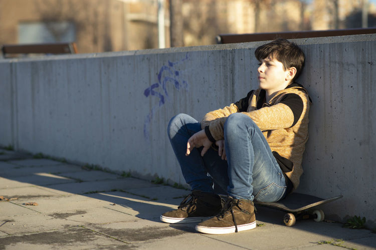 Young teenage sitting on his skateboard and leaning on fence
