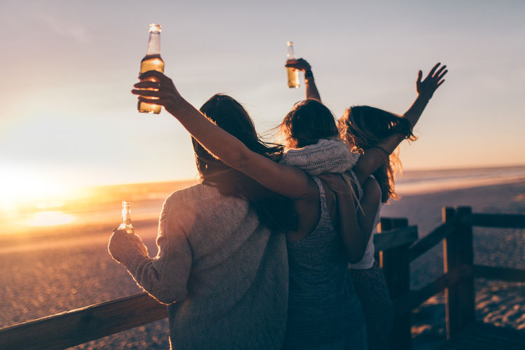 Women holding beer bottles while standing by railing against sea during sunset