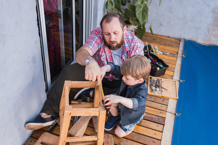 From above unshaven mature dad with attentive boy measuring wooden blocks with tape while spending time on blurred background