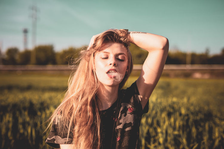 Beautiful young woman exhaling smoke on field against sky