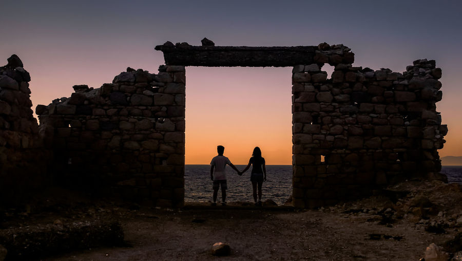Silhouette friends holding hands while standing at damaged wall during sunset