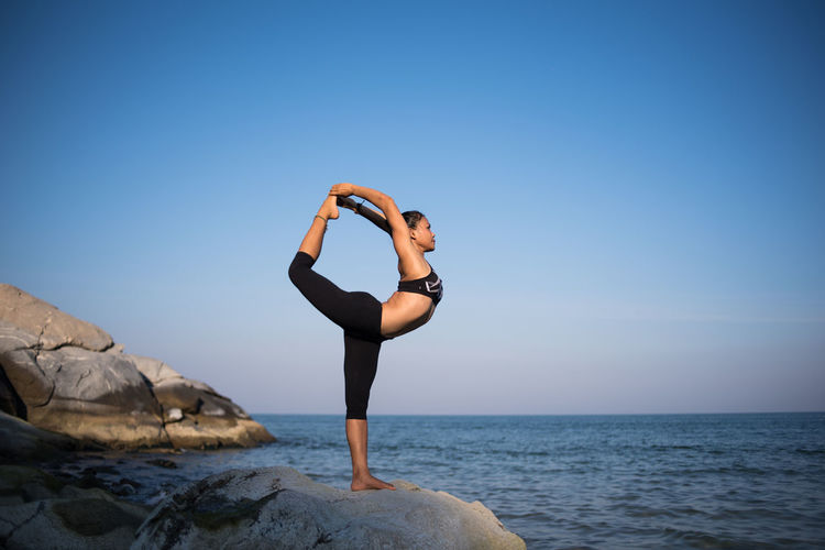 Side view of woman practicing natarajasana on rocky shore against blue sky
