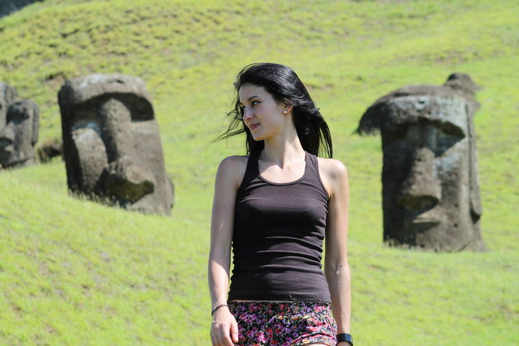 Young woman looking away while walking on grassy field against moai statue