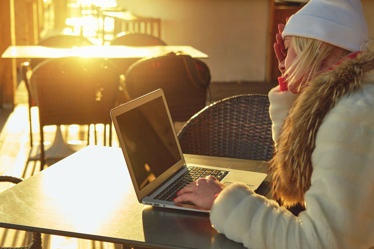 A woman in a sweater and a fur coat is sitting on the terrace of a cafe with a laptop.