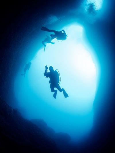 Unrecognizable professional divers with scuba and flippers swimming in narrow dark cave with rough stony formations undersea in deep clear water