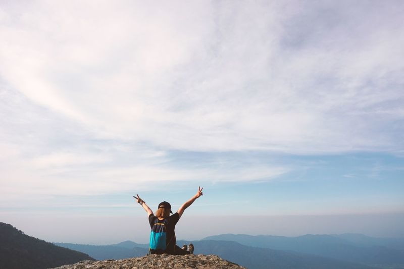 Woman gesturing peace sign while sitting on mountain against sky