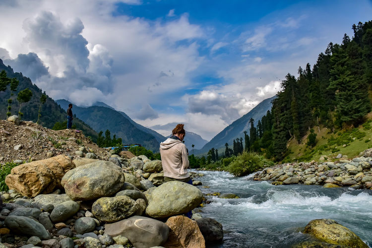 Rear view of woman sitting on rock by river against sky