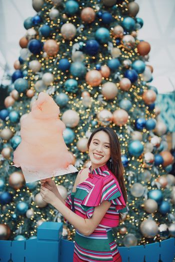 Portrait of smiling young woman standing by christmas tree holding cotton candy outdoors