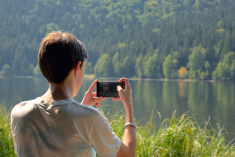 Rear view of teenage boy photographing while standing by lake