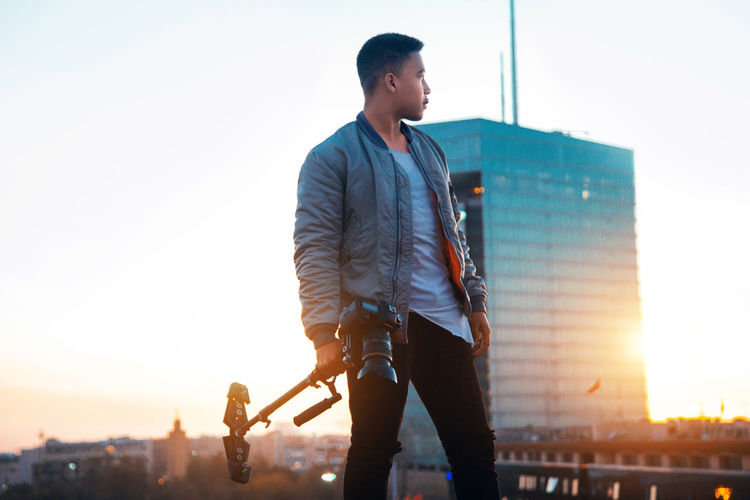 Young man holding camera while looking away in city against sky