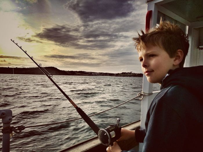 Side view of boy fishing in sea against sky during sunset