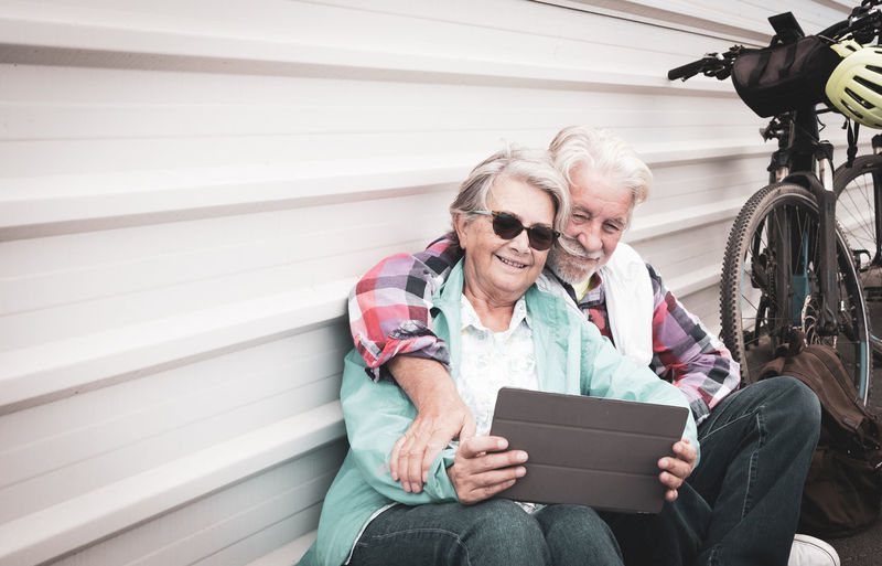 Smiling couple using digital tablet while sitting against wall