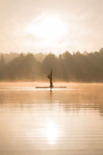 Silhouette person doing headstand in lake by trees against sky