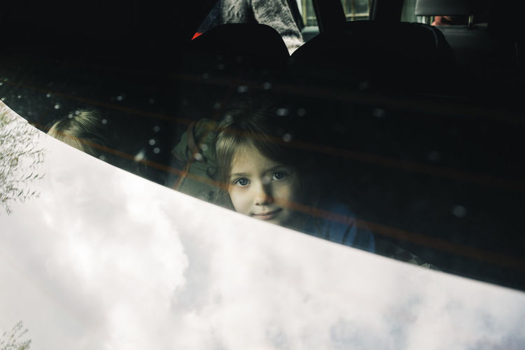 Portrait of girl sitting in electric car trunk seen through rear windshield