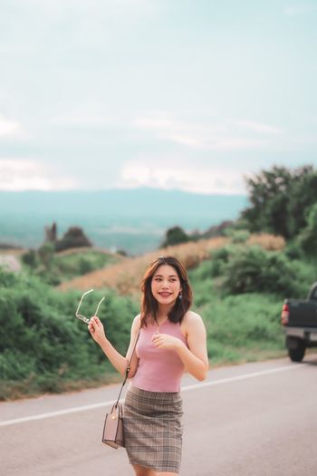 Portrait of smiling young woman standing on road against sky