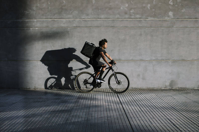 Side view of black delivery rider with box and bike riding on city pavement against wall with shadow while looking away in sunlight