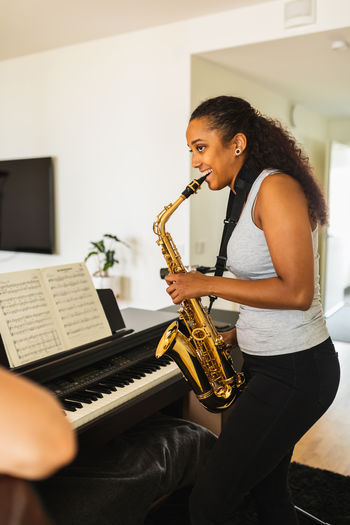 Side view of pensive hispanic female with long hair in casual clothes standing near piano with sheet music while plying saxophone in light room in apartment