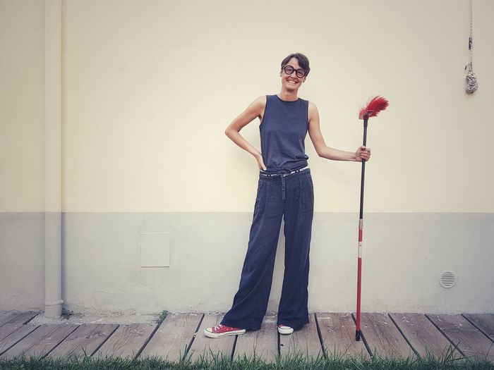 Portrait of smiling woman holding broom while standing against wall