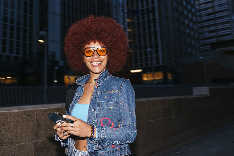 Attractive female with afro hairstyle and trendy outfit text messaging on cellphone while standing on street with modern buildings in evening time