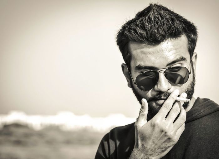 Close-up of fashionable young man wearing sunglasses smoking cigarette against sky