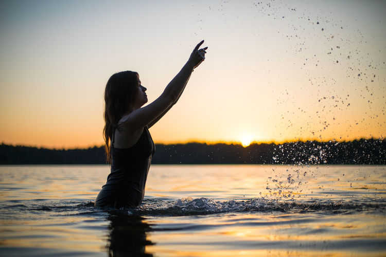 Silhouette woman with arms outstretched standing in lake during sunset
