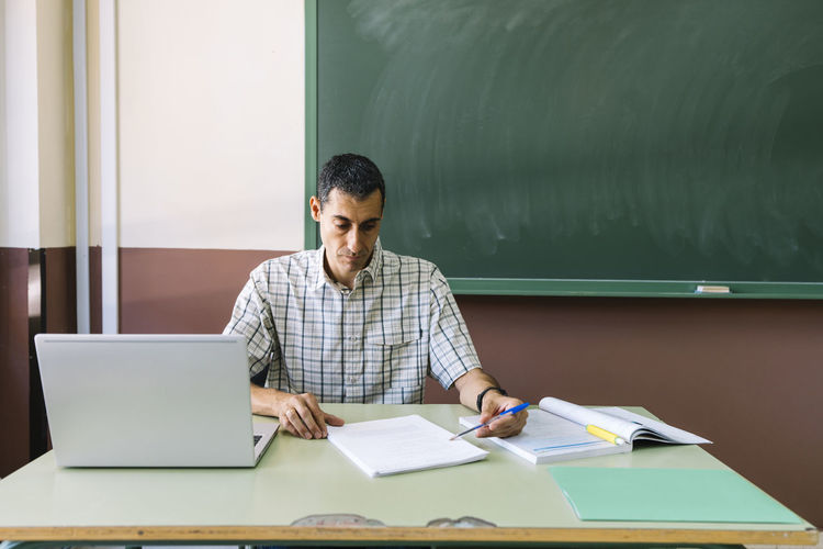 Professor sitting with laptop and checking paper in classroom