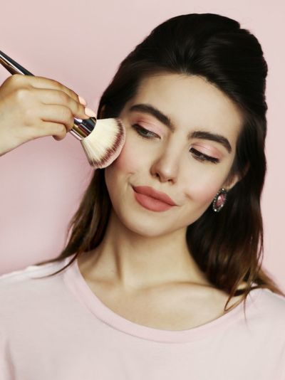 Close-up of beautiful young woman applying make-up on cheek against wall