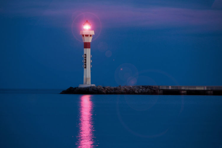 Illuminated lighthouse by sea against sky at dusk