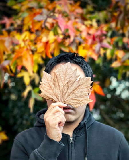 Portrait of man holding a dried autumn leaf against autumn foliage and trees.