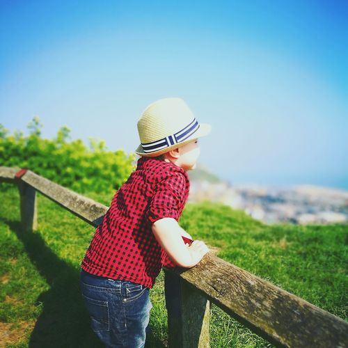 Side view of boy wearing hat standing by wooden railing against clear blue sky