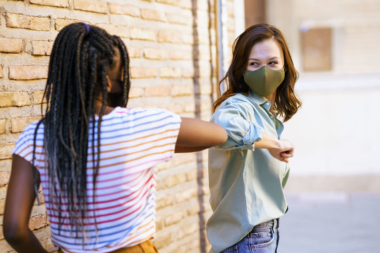 Smiling friends wearing mask doing elbow bump against wall