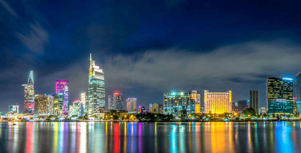 Illuminated urban skyline by river against sky during sunset