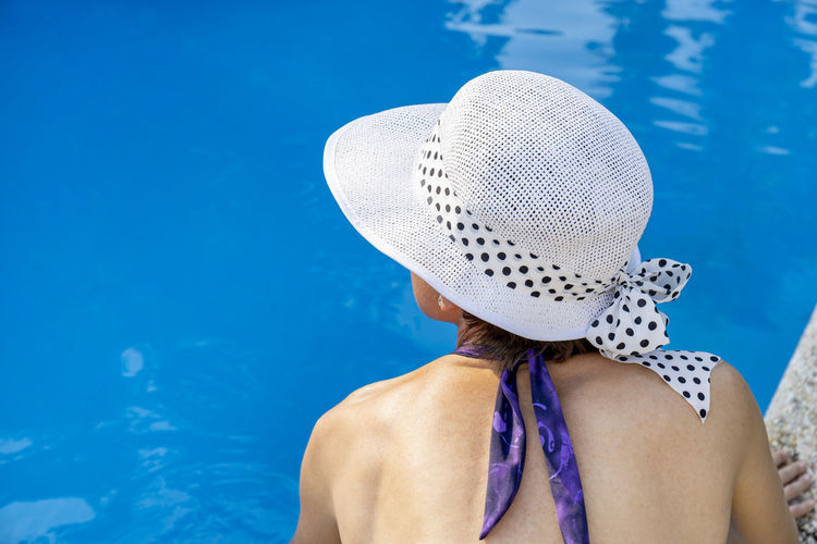A woman sitting on the edge of a swimming pool is wearing a purple bikini and a white hat.
