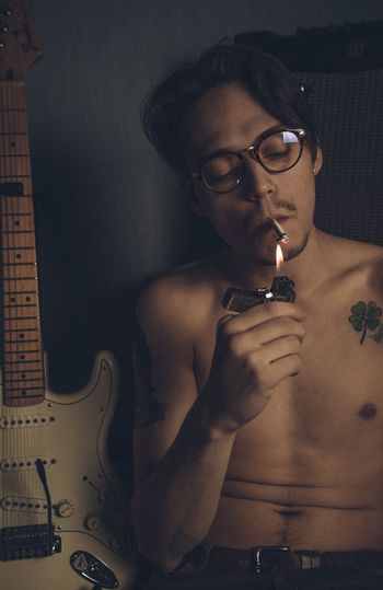 Shirtless young man igniting cigarette with lighter by guitar