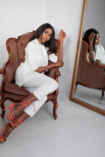 Full length portrait of african nigerian in white pants suit sitting in velvet brown chair in studio