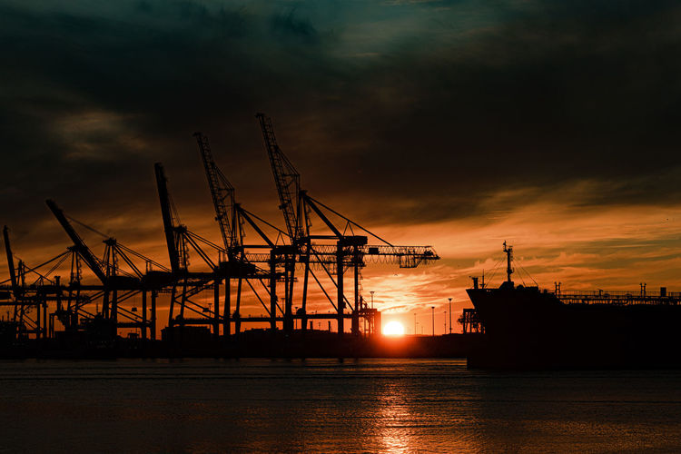 Silhouette cranes at harbor against sky during sunset